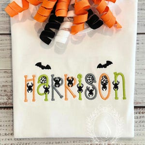 May include: A white t-shirt featuring the embroidered name Harrison in orange, lime green, and gray letters, decorated with black spider and web motifs. Two black bats are stitched above the name, and orange, black, and white ribbon curls are attached at the neckline.