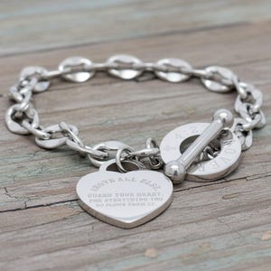 May include: A silver-colored bracelet with a chain-link design and a heart-shaped charm. The charm is engraved with the words "ABOVE ALL ELSE, GUARD YOUR HEART, FOR EVERYTHING YOU DO FLOWS FROM IT." The bracelet has a toggle clasp.