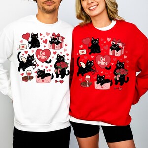 May include: White and red sweatshirts with a Valentine's Day theme. Each sweatshirt features a repeating pattern of black cat illustrations with hearts, gifts, and the text "Be Mine". The cats are depicted with wings, holding arrows, flowers, and balloons.