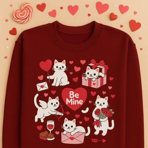 May include: Burgundy sweatshirt with a Valentine's Day theme. Features white cartoon cats with hearts, gifts, and Cupid's bow. The words "Be Mine" are on a heart. Decorative hearts and a heart-shaped lollipop are scattered around the sweatshirt.