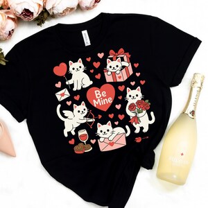 May include: Black t-shirt featuring a Valentine's Day design with white cartoon cats, red hearts, and the words "Be Mine." The cats are depicted with various Valentine's Day items, such as a balloon, a gift, and roses.