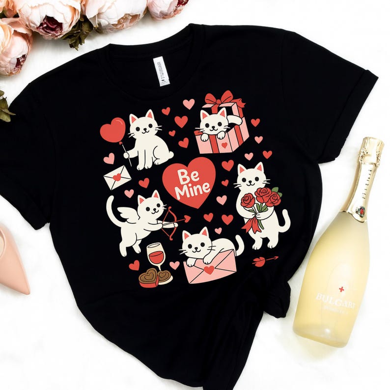 May include: Black t-shirt featuring a Valentine's Day design with white cartoon cats, red hearts, and the words "Be Mine." The cats are depicted with various Valentine's Day items, such as a balloon, a gift, and roses.