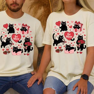 May include: Cream-colored t-shirts featuring a Valentine's Day design. The design includes black cat illustrations with hearts, gifts, roses, and the words "Be Mine". The shirts are worn by two people.