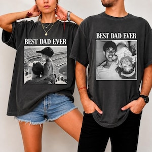 May include: Two dark gray t-shirts with the text "BEST DAD EVER" above a black and white photo. One shirt features a photo of a father and child at a baseball game. The other shirt shows a father and child smiling.