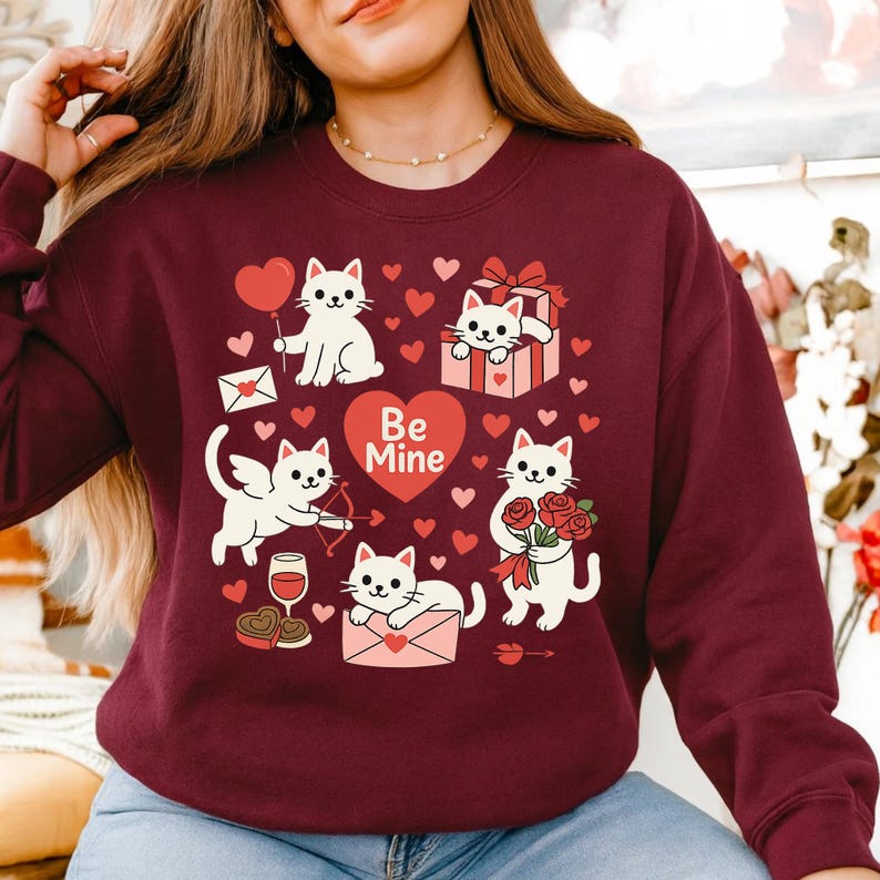May include: Burgundy sweatshirt featuring a Valentine's Day design with white cartoon cats holding a heart balloon, roses, and gifts. The phrase "Be Mine" is on a heart. Small red hearts are scattered around the cats.