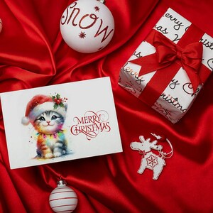 Könnte beinhalten: Eine weihnachtliche Szene mit einer Grußkarte mit einem Aquarellkätzchen mit Weihnachtsmütze und Lichterkette. Ein Geschenk mit roter Schleife, weiße Kugeln mit "snow"-Schriftzug und ein Rentier-Ornament liegen auf rotem Satin.