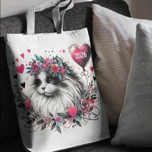Könnte beinhalten: Weiße Tragetasche mit einer schwarz-weißen Katzenillustration, die einen Blumenkranz trägt. Ein herzförmiger Ballon mit dem Jahr "2026" schwebt über der Katze. Rosa Herzen und florale Details schmücken das Design.