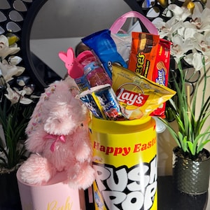 May include: A yellow Push Pop Candy container filled with snacks and candy, including Lay's chips and Reese's Peanut Butter Cups. A pink plush bunny and a pink box with the name "Riah" are also present, creating an Easter-themed gift.