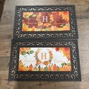 May include: Two rectangular doormats with a decorative border. Each mat features a central rectangular design with the text "Seasons Greetings" and the letter "H" surrounded by fall-themed imagery of pumpkins and leaves in shades of orange, yellow, and green.