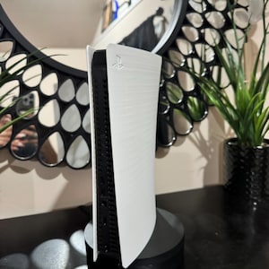 May include: A white and black gaming console standing upright on a black base. The console has a sleek, modern design with a curved white exterior and a black interior. The PlayStation logo is visible on the console.