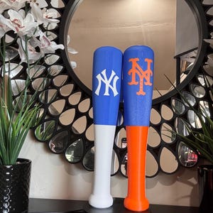 May include: Two baseball bats standing upright. The left bat is blue with a white New York Yankees logo. The right bat is blue with an orange New York Mets logo. The left bat has a white handle, and the right bat has an orange handle.