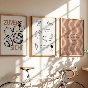 Könnte beinhalten: Drei gerahmte Kunstdrucke mit Holzrahmen hängen an einer Wand. Die Drucke zeigen Schwarz-Weiß-Illustrationen von Zitronen, Lebensmitteln und abstrakten Mustern. Der Text auf den Drucken ist auf Deutsch.