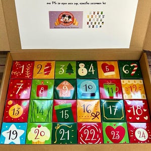 May include: Advent calendar box with 24 colorful, numbered compartments. Each compartment is decorated with festive images like a Christmas tree, snowman, and heart. The box includes text that says "ONE PIN TO OPEN EACH DAY, STARTING DECEMBER 1ST."