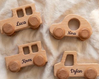 May include: Four wooden toy cars with names Luca, Nora, Knox, and Dylan engraved on them. The cars are light brown with wooden wheels. The cars are on a white, textured surface.