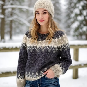 May include: A woman wearing a beige knitted hat and a patterned sweater in shades of grey, beige, and white. The sweater has a crew neck and long sleeves. The background is a snowy forest.