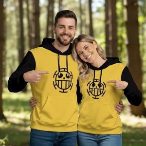 K&ouml;nnte beinhalten: Zwei Personen tragen passende gelbe Kapuzenpullover mit schwarzen &Auml;rmeln und einer schwarzen Grafik auf der Vorderseite. Die Grafik ist ein stilisierter Totenkopf mit Kreuz und Linien. Der Hintergrund ist ein verschwommener Wald.