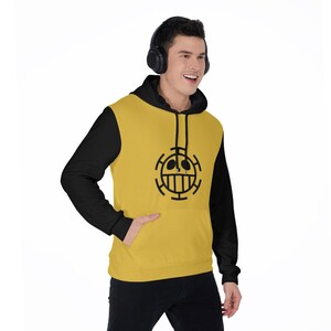 K&ouml;nnte beinhalten: Ein gelb-schwarzer Kapuzenpullover mit einer schwarzen Grafik auf der Vorderseite. Der Kapuzenpullover hat eine schwarze Kapuze und &Auml;rmel sowie eine K&auml;ngurutasche. Die Grafik ist ein schwarzes kreisf&ouml;rmiges Design mit einem Gesicht. Die Person tr&auml;gt eine schwarze Hose.