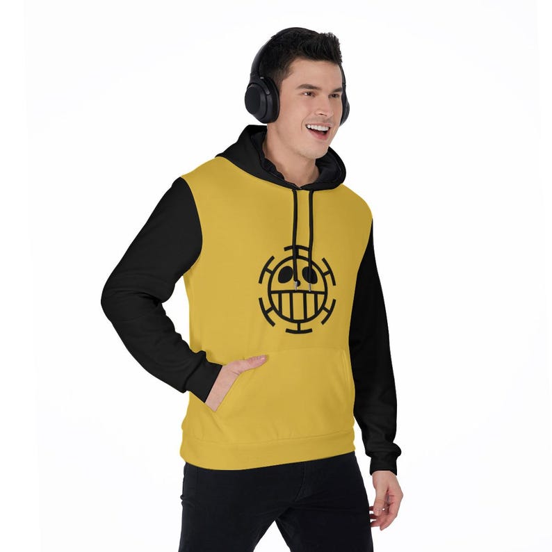 K&ouml;nnte beinhalten: Ein gelb-schwarzer Kapuzenpullover mit einer schwarzen Grafik auf der Vorderseite. Der Kapuzenpullover hat eine schwarze Kapuze und &Auml;rmel sowie eine K&auml;ngurutasche. Die Grafik ist ein schwarzes kreisf&ouml;rmiges Design mit einem Gesicht. Die Person tr&auml;gt eine schwarze Hose.