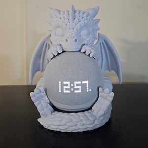 May include: A light gray dragon-shaped speaker holder cradling a gray smart speaker. The speaker displays the time, 12:57. The dragon has detailed wings, horns, and claws, creating a fantasy-themed decorative piece.