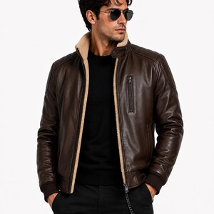 Puede incluir: Chaqueta de cuero marrón con cuello y forro de borreguillo beige. La chaqueta tiene cierre de cremallera, un pequeño bolsillo en el pecho y puños de canalé. La persona lleva pantalones negros y gafas de sol.