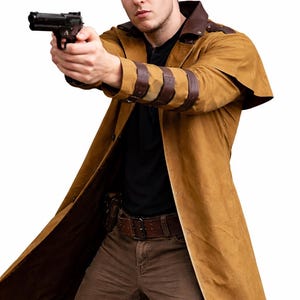 Puede incluir: Un abrigo largo color canela con detalles de cuero marrón en los hombros y las mangas. El abrigo está abierto, revelando una camisa negra y pantalones marrones. Se ve un cinturón y una funda de cuero marrón. Se sostiene una pistola negra.