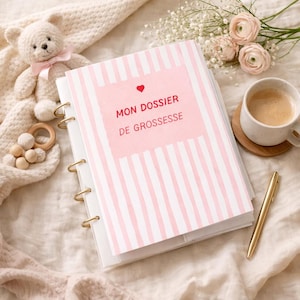 Pode incluir: Uma pasta listrada rosa e branca com o texto "MON DOSSIER DE GROSSESSE" em vermelho, um coração vermelho e anéis de metal dourado. Uma caneta dourada, uma xícara de café, um ursinho de pelúcia e um brinquedo de madeira também estão na imagem.