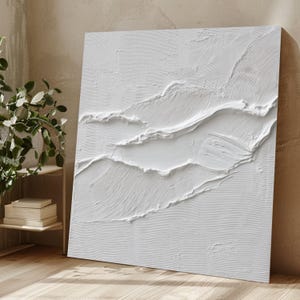 Puede incluir: Una gran obra de arte cuadrada y blanca texturizada con un diseño en capas en relieve. La obra de arte presenta una superficie texturizada con líneas horizontales y curvas, creando una sensación de profundidad y movimiento. La pieza está apoyada contra una pared de color neutro.
