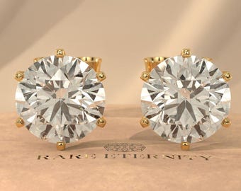 May include: A pair of gold stud earrings with large, round, clear gemstones. The earrings are set in a six-prong setting and rest on a pink surface atop a marble pedestal. The background is a soft, neutral color.