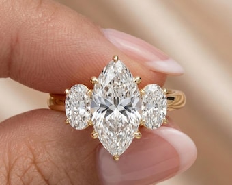 Pode incluir: Um anel de ouro com um diamante central em corte marquise e dois diamantes em corte oval em cada lado. O anel é segurado por dedos, destacando o brilho dos diamantes e o design.
