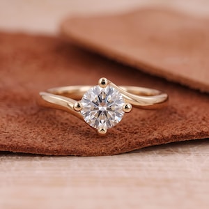 May include: A gold ring featuring a single round brilliant-cut clear gemstone held in a four-prong setting. The band has a smooth, polished finish and a bypass design that curves gently around the central stone, creating a sleek and continuous look.