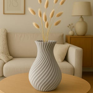 Könnte beinhalten: Eine graue, strukturierte Vase mit einem Spiralmuster enthält getrocknete, hellbraune Blumen. Die Vase steht auf einem runden, hellen Holztisch. Der Hintergrund umfasst ein beiges Sofa, eine Lampe und ein gerahmtes Kunstwerk.