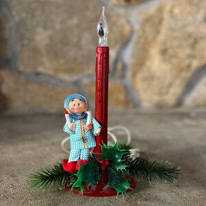 May include: A vintage Christmas light featuring a red candle with a light bulb at the top. A small, blue-clad figurine stands next to the candle, and green holly sprigs surround the base.