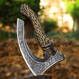 Puede incluir: Un hacha artesanal de hoja curva con un mango decorativo. El mango presenta un patrón texturizado dorado y negro. La hoja tiene intrincados diseños grabados y un símbolo en la base. El hacha se exhibe sobre una superficie de madera.