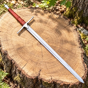May include: A longsword with a double-edged steel blade, a silver-toned metal crossguard, and a cylindrical grip wrapped in reddish-brown leather. The sword features a rounded pommel at the end of the handle and a tapered, pointed tip.