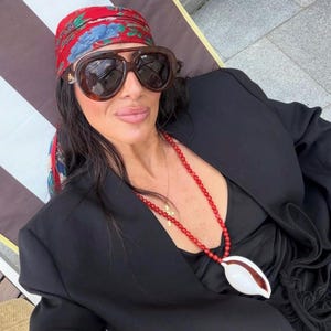 Puede incluir: Un pañuelo rojo con estampado floral, gafas de sol grandes de carey marrón y un collar largo de cuentas rojas pequeñas con un colgante de concha cauri blanca, combinado con una americana negra y una prenda superior negra.