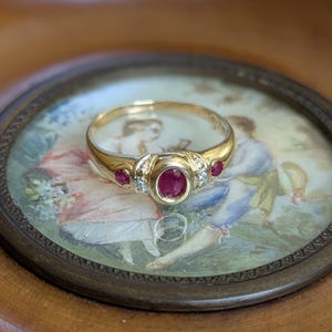 Può includere: Un anello d'oro con un rubino ovale centrale e rubini più piccoli su ogni lato. L'anello presenta anche piccole pietre di accento trasparenti. L'anello è posto su un piccolo dipinto rotondo incorniciato con una scena romantica.