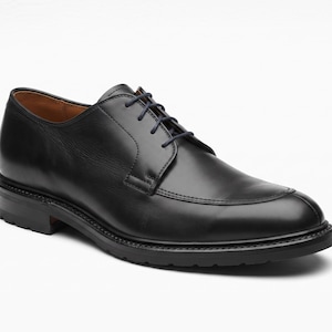 May include: A black leather derby shoe featuring a rounded toe, a stitched apron front, and a lace-up closure with thin black laces. The shoe has a low heel and a thick, textured black rubber sole, with a smooth leather interior lining visible.