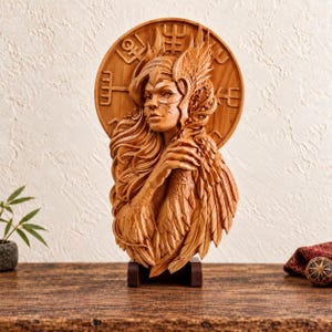 Op de afbeelding: Een lichtbruine houten wandplaat met een gedetailleerd reliëf van een gevleugelde figuur met lang, golvend haar. De figuur is geplaatst voor een cirkelvormige schijf die is gegraveerd met diverse symbolen. Het beeldhouwwerk rust op een donkere voet.