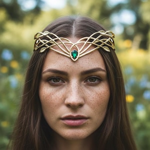 May include: A gold-colored headpiece with an intricate, interwoven design, featuring a central green gemstone. The headpiece has leaf-like accents and is worn on a person's head. The background is blurred, with green and yellow tones.