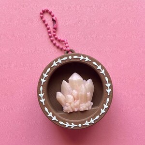 May include: A brown pendant with a white leaf design around the edge, holding a cluster of light pink crystals. A pink beaded chain is attached to the pendant. The background is a solid pink color.
