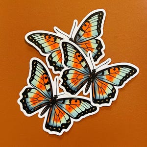 May include: Four butterfly stickers with orange, light blue, and black wing patterns. The stickers have a white border and are arranged on a brown surface. The butterflies have detailed wing patterns.