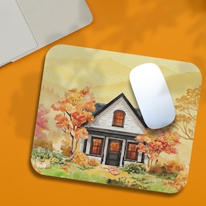 May include: A rectangular mousepad featuring a watercolor painting of a white house with a black roof and trim, surrounded by autumn trees with orange and red leaves. A white computer mouse rests on the pad, near a laptop.