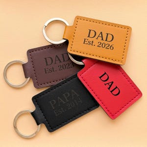 May include: Four rectangular leather keychains in yellow, dark brown, black, and red. Each features a silver metal ring and black engraved text. The yellow one reads DAD Est. 2026, the brown one DAD Est. 2023, the black one PAPA Est. 2014, and the red one DAD.