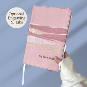 Peut inclure: Un carnet rose avec un motif de paysage abstrait texturé dans des tons de rose, blanc et doré. La couverture porte le texte "my dear, Kayla" et un marque-page en ruban blanc. Un graphique circulaire affiche le texte "Optional Engraving & Tabs".