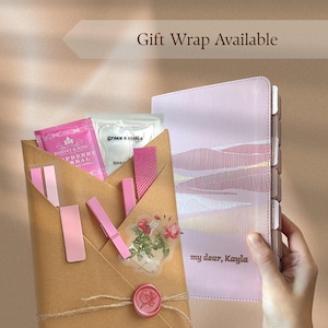 Peut inclure: Un agenda rose clair avec un motif de paysage et le texte 'my dear, Kayla' à côté d'une enveloppe en papier kraft contenant un sachet de thé Harney & Sons, un masque pour les yeux Grace & Stella, des pinces roses et un autocollant floral.