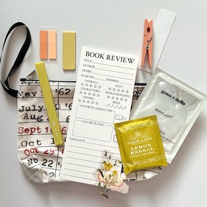 May include: A flat lay of book review supplies, including a canvas tote bag with calendar-themed print, a book review sheet, sticky notes, a highlighter, a tea bag, and eye masks. A wooden clothespin and a nail file are also included.