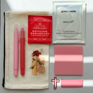 May include: A flat lay of beauty and self-care items. Includes a canvas pouch, two lip pencils, a Harney & Sons English Breakfast tea bag, a packet of Grace & Stella Firming Eye Masks, a pink notepad, a paper clip, and a small pink rectangular object.