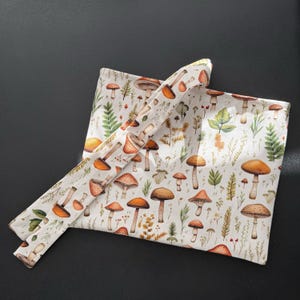 May include: A square fabric wrap featuring a repeating pattern of various brown and orange mushrooms interspersed with green ferns and small yellow wildflowers on a white background. The fabric has a long, narrow tie attached to one corner.