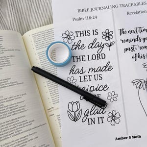 May include: An open Bible next to Bible journaling traceables with the text "This is the day the Lord has made, let us rejoice and be glad in it." A black pen and blue washi tape are also present.