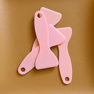 May include: Several light pink silicone spatulas are arranged on a brown surface. Each spatula has a flat, angled blade and a long handle with a hole for hanging. The spatulas vary slightly in shape, with some having rounded or pointed blades.